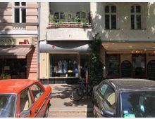 Eine Straßenszene mit einem kleinen Geschäft mit Schaufenster in der Mitte, flankiert von einem Buchladen links und einem Restaurant mit Tafeln rechts. Davor parken ein oranger Oldtimer und ein schwarzes Auto, daneben steht ein Fahrrad an der Hauswand.