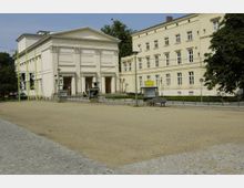 Historisches Theatergebäude mit klassizistischer Fassade, auf der "Maxim Gorki Theater" steht, daneben ein benachbartes, schlichtes Gebäude mit mehreren Fenstern. Im Vordergrund ein offener Platz mit Kiesboden, Sitzbänken und Infotafeln.