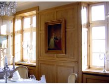 Holzvertäfelter Speiseraum mit großen Fenstern, die viel Licht hereinlassen, einem gerahmten Gemälde an der Wand und einem festlich gedeckten Tisch mit Kerzenleuchtern und gefalteten Servietten.