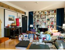 Wohnzimmer mit Büroecke und Bücherregal: Im Vordergrund ein Schreibtisch mit Dokumenten, Fotos und Schreibutensilien. Im Hintergrund ein großes weißes Bücherregal, farbige Möbel, Dekoration sowie ein schwarzes Sideboard mit darüber hängenden gerahmten Bildern.