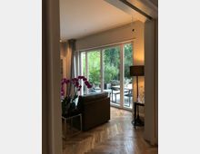 Wohnzimmer mit Parkettboden und großer Fensterfront, die auf eine Terrasse mit Gartenblick führt. Im Raum befinden sich eine Couch, ein Tisch mit Orchideen und eine Stehlampe, die eine gemütliche Atmosphäre schafft.