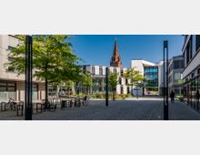 Moderner Innenhof mit gepflastertem Boden, umgeben von Bürogebäuden mit großen Glas- und Putzfassaden. Im Vordergrund Bäume, Fahrradständer und schlanke schwarze Laternen, im Hintergrund der Turm einer roten Backsteinkirche unter blauem Himmel.