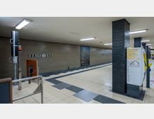 U-Bahn-Station mit der Bezeichnung „Britz-Süd“, gekennzeichnet durch graue Wandfliesen, schwarze Säulen und gelb-weiße Ticketautomaten. Der Boden ist mit hellen Fliesen und taktilen Leitstreifen ausgestattet, und an einer Wand ist ein Bild eines blühenden Baumes zu sehen.