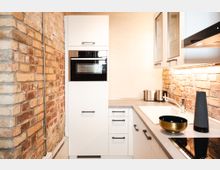 Moderne Küche mit weißer Einbauküche, grauer Arbeitsplatte und Backofen in einem Hochschrank. Die Wände sind teils mit Sichtmauerwerk aus roten Ziegelsteinen gestaltet, und die Beleuchtung unter den Hängeschränken hebt die Arbeitsfläche hervor.