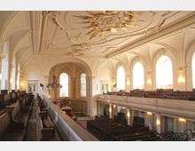 Innenraum einer barocken Kirche mit weißen Wänden, hohen Rundbogenfenstern und reich verzierten Decken mit goldenen Ornamenten. Im Vordergrund sind Holzbänke auf einer Empore, im Hintergrund der Altarraum mit großen Fenstern und prächtiger Kanzel.