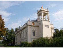Klassizistisches Gebäude mit beigefarbener Fassade, Flachdach und einem Säulenturm, der die deutsche Fahne trägt. Das Gebäude ist von Bäumen und Büschen umgeben und steht unter einem hellblauen Himmel.
