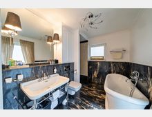 Modernes Badezimmer mit Marmorböden und -wänden in Schwarz mit goldenen Akzenten. Zu sehen sind ein freistehendes weißes Badewannenmodell, ein großer Spiegel mit Wandlampen, ein Waschbecken mit Chromarmaturen und dekorativen Accessoires, sowie ein kleines Fenster mit beigefarbenem Rollo.