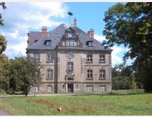 Historisches, dreigeschossiges Gebäude mit symmetrischer Fassade, Schieferdach und einem Storchenhorst auf dem Dach. Es ist umgeben von einer Grünfläche mit Bäumen und befindet sich in einer ländlichen Umgebung.