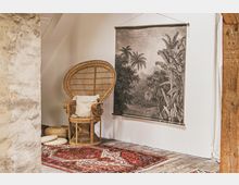 Gemütlich gestalteter Raum mit einer Rattan-Sitzgelegenheit, einem cremefarbenen Kissen und einem dekorativen Wandteppich mit tropischem Motiv. Der Boden ist mit einem roten, orientalischen Teppich bedeckt, und im Vordergrund befindet sich eine raue Wand mit sichtbarer Struktur.