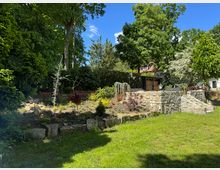 Gepflegter Gartenbereich mit einer niedrigen Steinmauer und Treppe aus Naturstein, umgeben von grüner Rasenfläche, Blumenbeeten und hohen Bäumen. Im Hintergrund sind dichter Buschbewuchs, kleine Gebäude sowie ein Pavillon sichtbar.
