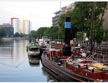 Hafenansicht an einem Fluss mit angedockten Booten und Schiffen, darunter ein größeres rotes Dampfschiff im Vordergrund. Im Hintergrund sind moderne Wohnhochhäuser in Weiß mit farbigen Akzenten sowie grüne Bäume und einflussnahe Gebäude sichtbar.