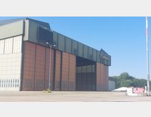 Ein großes Flugzeughangar-Gebäude mit einer grünen und orangefarbenen Metallfassade und geöffneten Toren, das mit dem Logo der Lufthansa gekennzeichnet ist. Im Vordergrund befindet sich ein leerer Asphaltbereich, im Hintergrund sind Bäume und ein kleinerer weißer Baucontainer sichtbar.