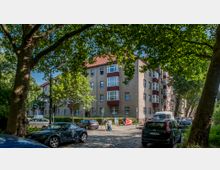 Mehrstöckiges Wohngebäude mit heller Fassade und roten Fensterrahmen, umgeben von Bäumen und parkenden Autos auf einer gepflasterten Straße. Im Vordergrund führt eine Person mit einem Kinderwagen einen Gehweg entlang.