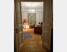 Blick in ein Arbeitszimmer mit Holzboden, einem großen Schreibtisch aus Holz, einem modernen Gemälde an der weißen Wand und einer hängenden Deckenleuchte. Links ein stehender Lampenschirm, auf dem Boden ein länglicher Teppich.