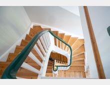 Blick von oben in ein helles Treppenhaus mit gewendelten Holzstufen und weißen Geländern. Ein grüner Handlauf führt entlang der Treppe, umgeben von weißen Wänden und Holzdetails.