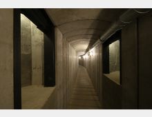 Betonierter, schmaler Tunnel mit leicht gewölbter Decke, seitlichen Fensteröffnungen und einer Metallrohrleitung an der rechten Wand. Der Boden besteht aus Holzplanken, und die Beleuchtung erfolgt durch eine Reihe von Glühbirnen entlang der Decke.