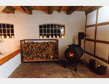 Gemütlicher Innenraum mit einem gemauerten Boden, weiß verputzten Wänden und Fachwerkelementen. Im Vordergrund ein Holzofen mit glühendem Feuer, daneben ein Korb mit Holzscheiten; gegenüber ein Regal mit gestapeltem Brennholz, darüber zwei Fenster mit Kerzen.