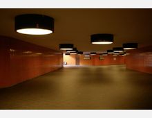 Unterführung mit orange gefliesten Wänden, schwarzem Boden und großen runden Deckenleuchten. Im Hintergrund sind Ausgänge und Wandtafeln erkennbar.