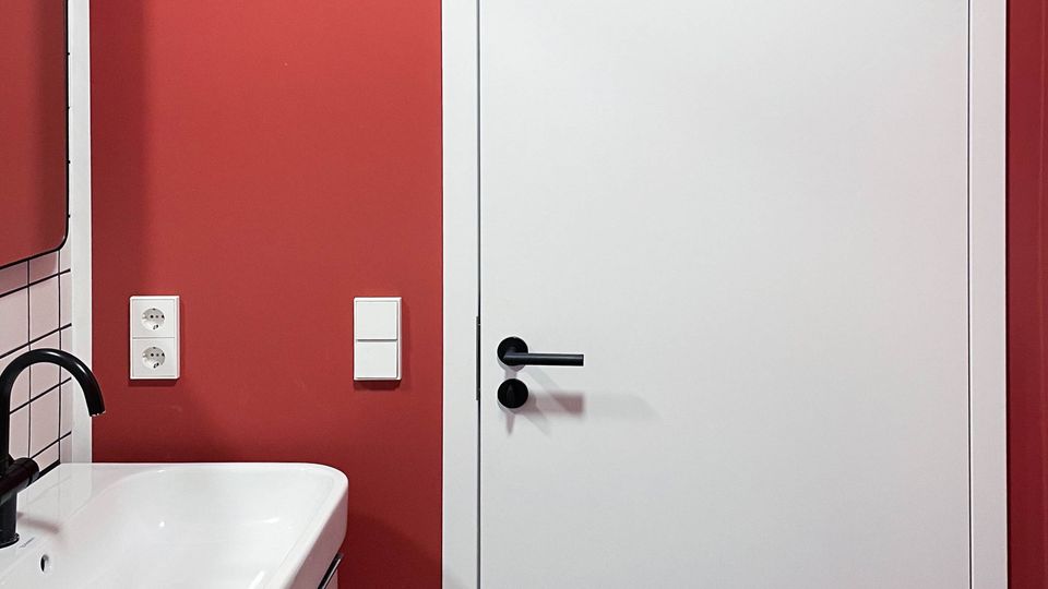 Badezimmer mit einer roten Wand, einer weißen Tür mit schwarzem Türgriff, einem weißen Waschbecken mit schwarzer Armatur auf der linken Seite und einem Boden mit schwarz-weißem Marmormuster.