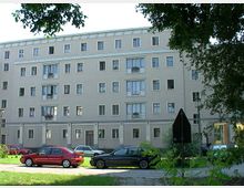 Wohngebäude mit vier Stockwerken und beigefarbener Fassade, mehreren Fenstern und Balkonen. Im Vordergrund stehen geparkte Autos, und Bäume rahmen das Bild ein.