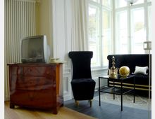 Wohnzimmer mit klassischem Design: Ein antiker Holzschrank mit einem Röhrenfernseher steht links neben einer schwarzen, hochlehnigen Samt-Sesselgarnitur. Große weiße Fenster lassen viel Tageslicht herein, und ein kleiner Glastisch mit dekorativen goldenen Objekten steht im Vordergrund.