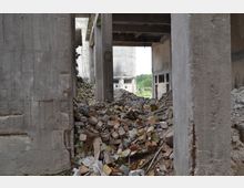 Innenansicht einer verlassenen, teils eingestürzten Betonruine mit beschädigten Wänden und freiliegenden Stahlträgern. Am Boden sind große Mengen an Ziegel- und Betonschutt zu sehen, im Hintergrund erstreckt sich eine grüne Landschaft durch zerstörte Fensteröffnungen.