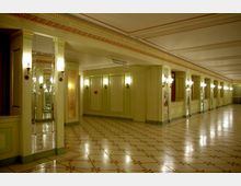 Innenansicht eines prunkvollen Foyers mit grünlich-gelben Wänden, Spiegeln, Säulen und symmetrisch angeordneten Wandleuchten. Der Boden ist mit glänzenden Fliesen im geometrischen Muster gestaltet, und die Decke ist mit Zierleisten und Ornamentbändern verziert.