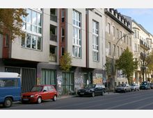 Straßenansicht eines modernen Wohngebäudes mit großen Fenstern und Balkonen, flankiert von älteren Gebäuden. Vor dem Gebäude sind geparkte Autos, ein Gehweg mit Bäumen und teils Graffiti an den unteren Fassaden erkennbar.
