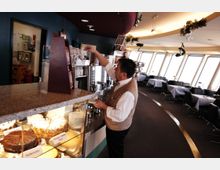 Ein Kellner in einer eleganten Weste steht hinter einer Theke in einem Café oder Restaurant. Im Vordergrund sind Torten in einer verglasten Vitrine zu sehen, während der Hintergrund eine modern gestaltete Speisehalle mit gedeckten Tischen und großen Fensterfronten zeigt.