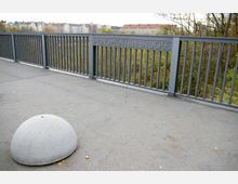 Eine Brücke mit einem grauen Metallgeländer, auf dem "Monumentenbrücke" steht, und einem halbkreisförmigen Betonelement auf dem Gehweg. Im Hintergrund sind Bäume und eine Häuserreihe zu sehen.