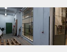 Industriehalle mit heller Beleuchtung, einer grünen Tür an der weißen Wand und einer großen Glaswand mit schwarzen Metallrahmen, die in einen angrenzenden Arbeitsbereich blicken lässt. Im Vordergrund ist eine Holzpalette, an den Wänden hängen Werkzeuge und Elektroinstallationen.