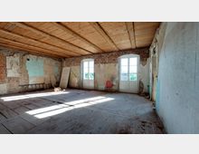 Ein leerer Raum mit sichtbarer Renovierungsbedürftigkeit, Holzdecke, abgenutztem Holzboden und Wänden aus freiliegendem Mauerwerk sowie verblassender Farbschicht. Zwei hohe Fenster mit weißen Rahmen lassen Tageslicht herein, während im Raum eine Leiter, ein Stuhl und ein Türblatt angelehnt stehen.