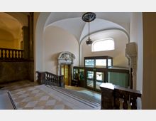 Treppenhaus eines historischen Gebäudeinneren mit breiten Marmorstufen, verzierten dunklen Geländern und einem großen bogenförmigen Fenster, das Tageslicht hereinscheint. Im Hintergrund befindet sich eine moderne Glasliftanlage und eine kunstvoll gestaltete Tür mit steinernem Dekor.