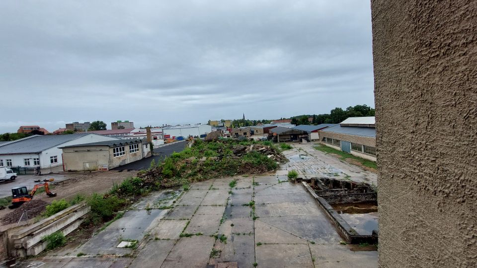 Industrie- oder Gewerbegebiet mit verfallenen Betonflächen, bewachsenen Erdaufschüttungen und mehreren Lagerhallen; im Hintergrund sind Siedlungsgebäude und ein Kirchturm sichtbar. Der Himmel ist bewölkt.