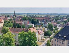 Luftaufnahme einer Kleinstadt mit roten Ziegeldächern und von Bäumen gesäumten Straßen; im Hintergrund ragt ein Kirchturm empor, umgeben von flachen Feldern und einer weitläufigen Landschaft.