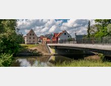 Eine Flusslandschaft mit einer modernen Betonbrücke im Vordergrund, die mit einem Geländer ausgestattet ist. Im Hintergrund steht eine Reihe von bunten Häusern mit Giebeldächern, umgeben von Bäumen und einer grünen Wiese am Ufer.