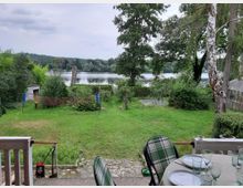 Ein Garten mit Rasenfläche, umgeben von Bäumen und Büschen, führt zu einem See im Hintergrund. Im Vordergrund befindet sich ein gedeckter Tisch auf einer Terrasse, und ein Badmintonnetz steht in der Mitte des Gartens.