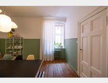 Helles Zimmer mit Holzdielenboden, einer weißen Wand im oberen Bereich und grün gestrichenen Wänden im unteren Bereich. Im Raum stehen ein Regal mit Geschirr, ein Tisch mit Stühlen und ein Fenster mit weißen Vorhängen, das Tageslicht hereinlässt.