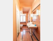 Langes Badezimmer mit rosafarbenen Fliesen, schwarzer Bodenbordüre und einem weißen Waschtisch mit großem Wandspiegel und rechteckigen Wandleuchten. Am Ende des Raums stehen ein WC und ein Fenster, daneben ein Handtuchheizkörper.