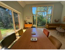 Raum mit einer großen Fensterfront, die den Blick auf einen Garten mit Bäumen und Sträuchern freigibt. Im Vordergrund steht ein Holztisch mit verschiedenfarbigen Stühlen, im Hintergrund eine gelbe Eckcouch, ein kleiner runder Couchtisch und eine Holzkommode.