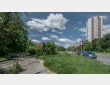 Eine städtische Landschaft mit einer asphaltierten Geh- und Fahrradwegauffahrt, umgeben von dichtem Grün und Bäumen links, einer Hauptstraße mit geparkten Autos und Ampeln rechts sowie einem hohen Wohngebäude im Hintergrund. Der Himmel ist klar mit einigen weißen Wolken.