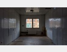 Leerer, schmaler Raum mit grauen, gefliesten Wänden und einer rechteckigen Deckenleuchte. An der Stirnseite befindet sich ein Fenster mit Gitterstäben, darunter ein Heizkörper, und die Wände zeigen sichtbare Gebrauchsspuren.