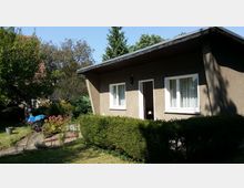 Kleines Wohnhaus mit Flachdach, verputzten braunen Wänden und weißen Fenstern. Im Vordergrund ein Garten mit Hecke, Blumenbeeten und einem Rasenbereich, umgeben von Bäumen und Sträuchern.