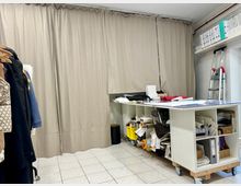 Ein Arbeitsraum mit einem langen Tisch auf Rollen, der mit Stoffproben, Papierrollen und Materialien belegt ist. Im Hintergrund befinden sich beige Vorhänge, Regale mit Ordnern und eine Anstellleiter, während links Kleidungsstücke auf Kleiderbügeln hängen.