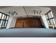 Großer, leerer Saal mit hoher, geometrisch gegliederter Decke und mehreren runden Deckenleuchten. An beiden Seiten verlaufen raumhohe Fensterfronten; die Stirnwand ist mit Holzvertäfelung und Türen gestaltet.