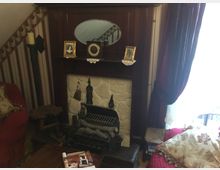 Ein traditionelles Wohnzimmer mit einem Kamin aus Holz und Stein, dekoriert mit einem ovalen Spiegel, gerahmten Fotos und einer kleinen Uhr. Im Vordergrund befinden sich ein rotes Sofa mit gemusterten Kissen und eine hölzerne Beistelltischgruppe, während von der rechten Seite Tageslicht durch einen Vorhang einfällt.