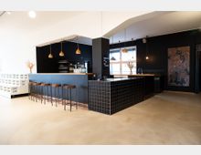 Moderne Bar mit schwarzer Fliesenverkleidung und Sitzhockern aus Holz. Helle Hängeleuchten, große Fenster, minimalistische Dekoration mit Trockenblumen und ein abstraktes Wandbild sind sichtbar, die den Raum definieren.