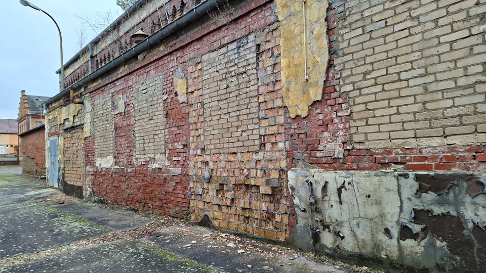 Seitenansicht eines alten Backsteingebäudes mit stark verwitterter Fassade, abgeplatztem Putz und teils ausgebesserten Mauerflächen. Entlang der Wand verläuft ein schmaler Gehweg; am Rand stehen eine Straßenlampe und im Hintergrund weitere Gebäude.