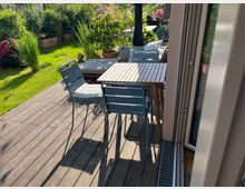 Holzterrasse mit blau-grauen Metallstühlen und einem Holztisch, umgeben von einem gepflegten Garten mit grünem Rasen, hohen Gräsern und bunt blühenden Pflanzen. Im Hintergrund ist eine Mischung aus Topfpflanzen und Gartendekorationen zu sehen.