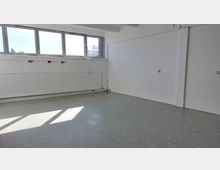 Ein leerer Raum mit weiß gestrichenen Wänden, grauem Linoleumboden und großen, rechteckigen Fenstern an der oberen Wand, die Tageslicht hereinlassen. Ein Heizkörper unter den Fenstern sowie sichtbare Rohrleitungen und Steckdosen sind an den Wänden angebracht.